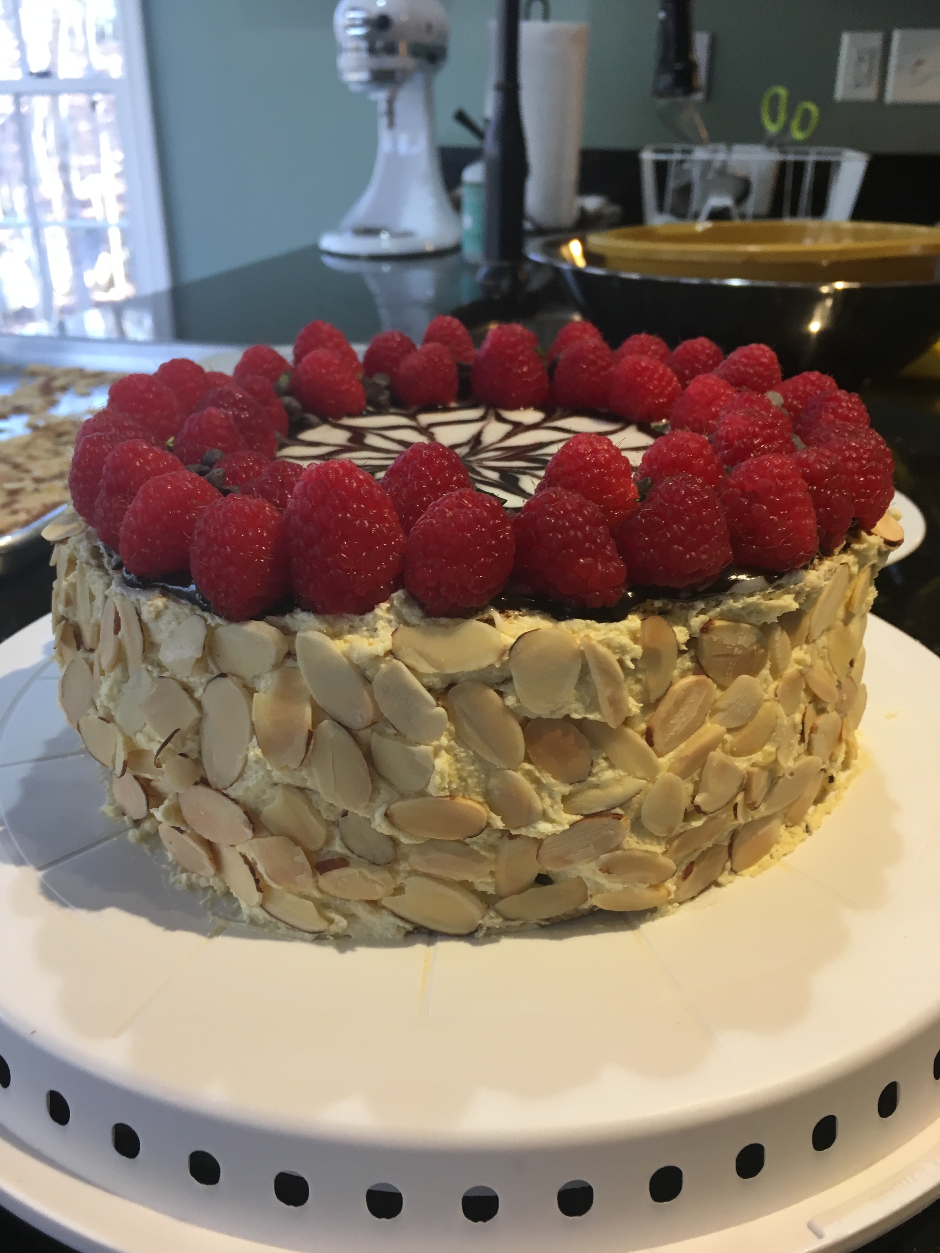 Finished Esterhazy cake with almonds covered on side, chocolate swirl flower on top, and raspberries in a circle on the top.